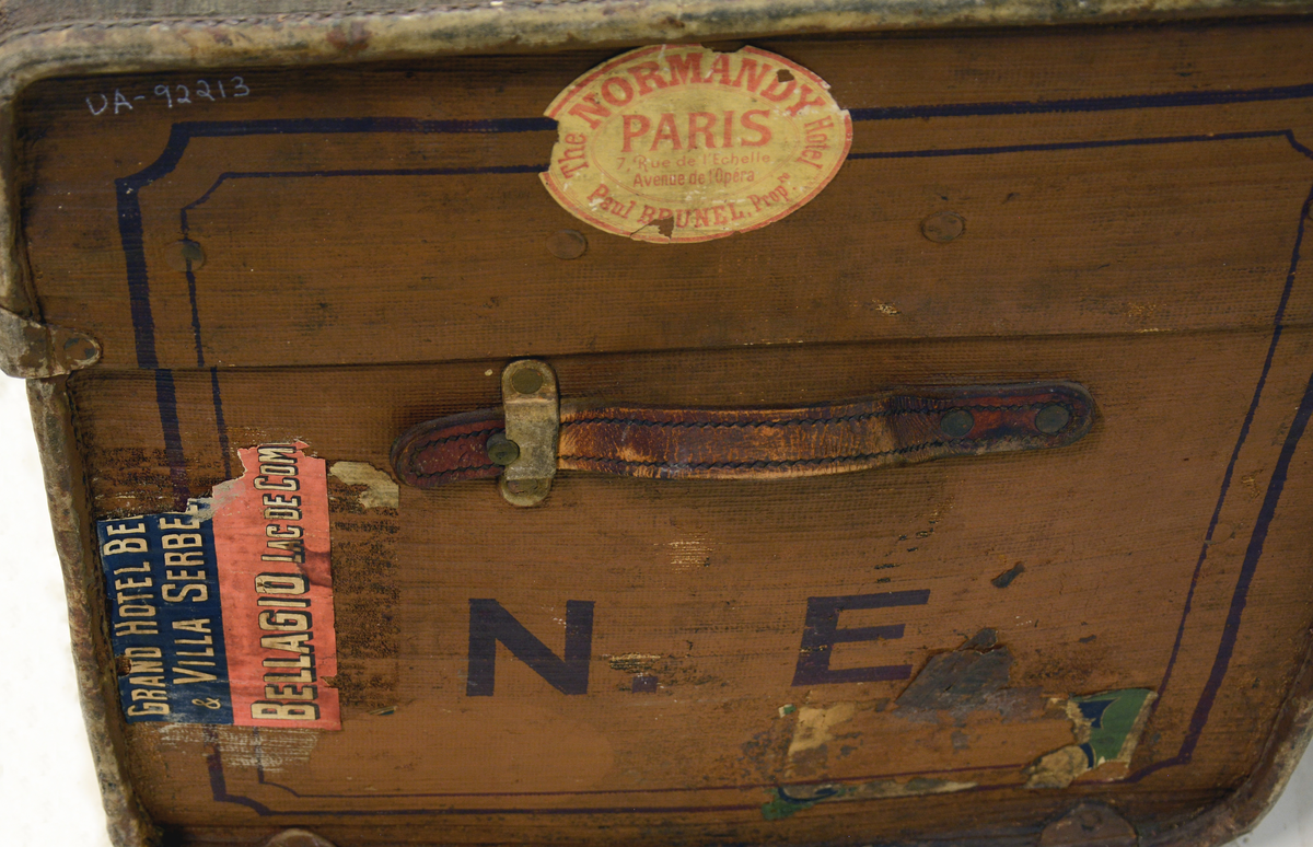 Koffert med flatt lokk. Etiketter pålimt fra ulike hoteller i bl.a. Paris og Kiel.
