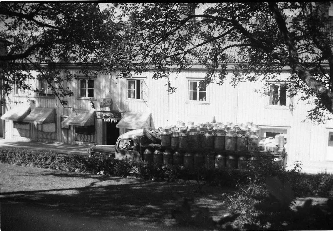 En fastighet på Brahegatan i Gränna, fotograferat mellan träd på motsatta sidan. Skylt för Folke Palms garn- och manufakturaffär. Markiserna är nedfällda på gatuvåningen. En mjölkbil passerar.