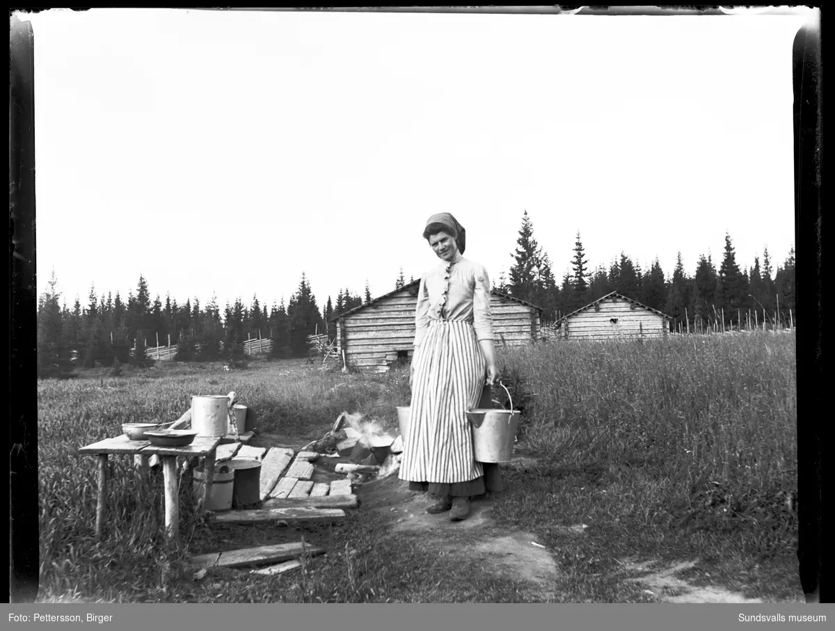 Fäbodbild från Klackbodarna i Stöde. Emma Flemström med två mjölkhinkar. I bakgrunden två timrade fäbodstugor och gärdesgård.