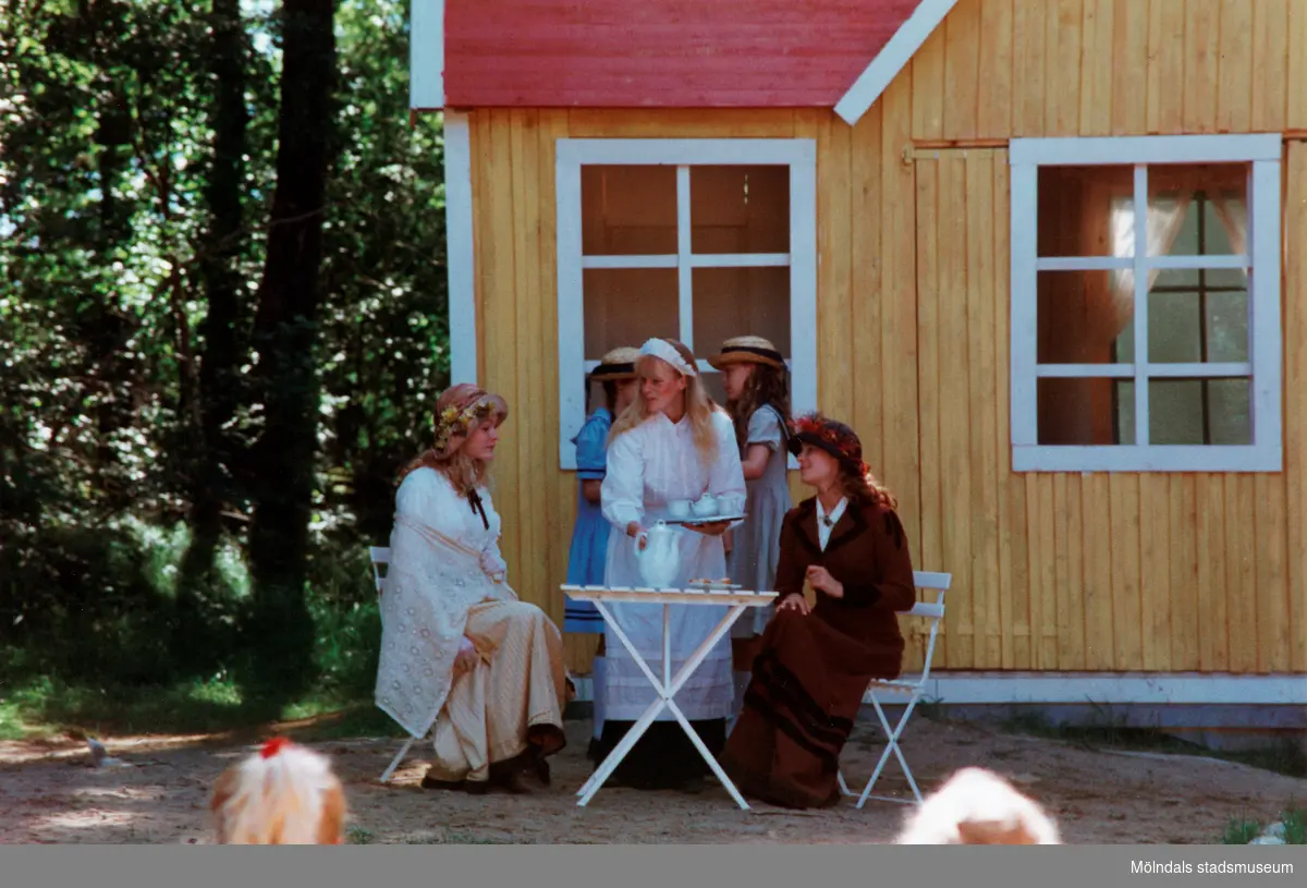 Sommarspelet/barnteaterföreställningen "Rasmus på luffen" 1999 vid Byxorna i Gunneboskogen (korsningen Christina Halls väg/John Halls väg). Föreställningen pågår och publiken ser på. Man ser två kvinnor som sitter vid ett kafé-bord framför ett gult litet hus. En kvinna serverar dem och i bakgrunden står två ungdomar.