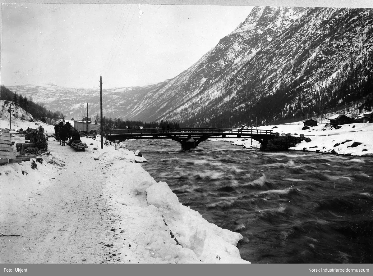 Bro på Såheim før industrialiseringen og bebyggelsen på Rjukan. To arbeidshester med slede stående på vei. Gårdsbebyggelse både på sør og nordsiden av elva Måna.