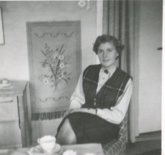 En kvinna sitter på en stol i ett rum, Backen eller Högen 1960-tal. Snett bakom henne hänger en bonad på väggen. Namnuppgift saknas.