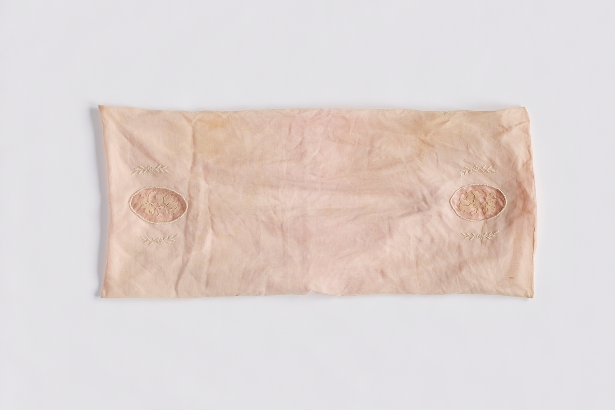 Et rektangulært rosa putetrekk sydd i to lag. Det innerste laget er av silke og det ytre laget er av silkechiffon. Det er åpning på undersiden for å tre inn pute. På oversiden er det broderinger og blonder i blomstermønster.