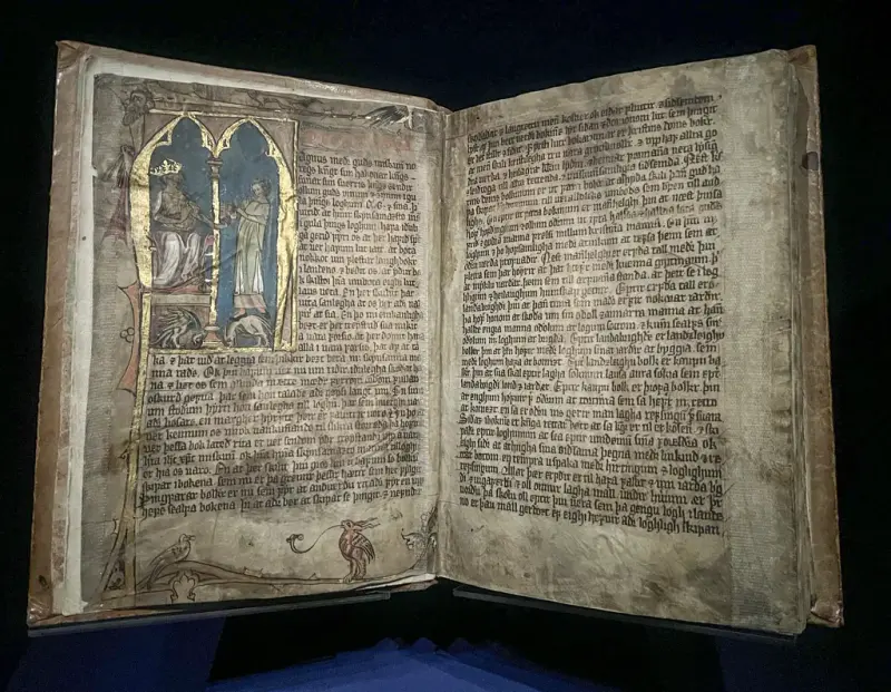 Middelalderpergament som viser to sider pergament med sirlig skrift og en illuminasjon på venstre side med motiv av to personer mot blå bakgrunn og gullramme rundt.