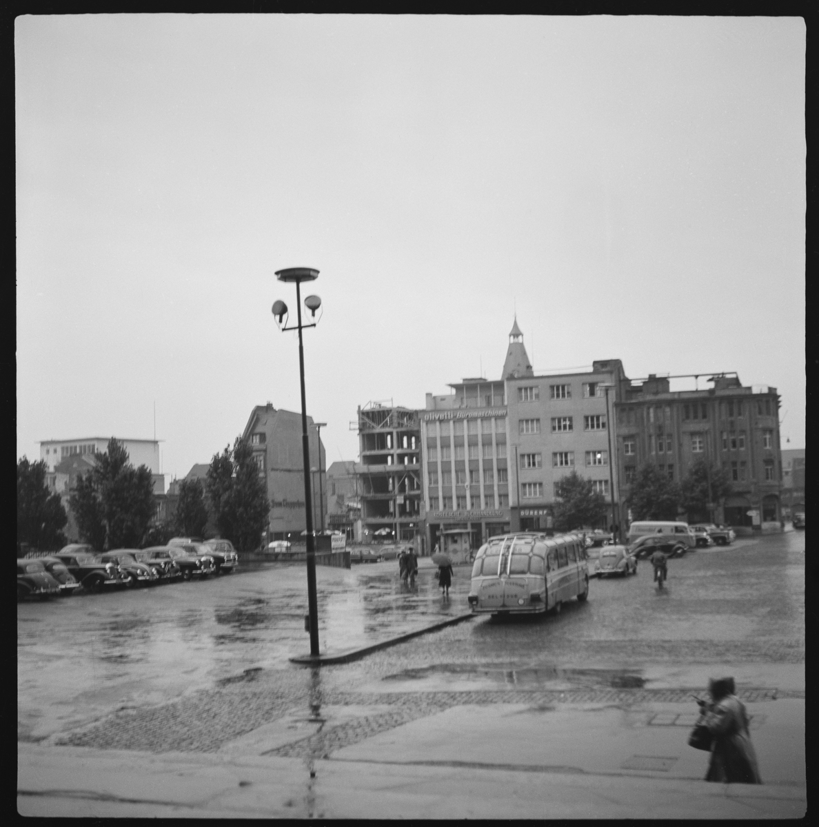 Et torg i en by i Tyskland en regnfull dag. Parkerte biler og en buss. Personer under paraplyer og med regntøy går over torget.