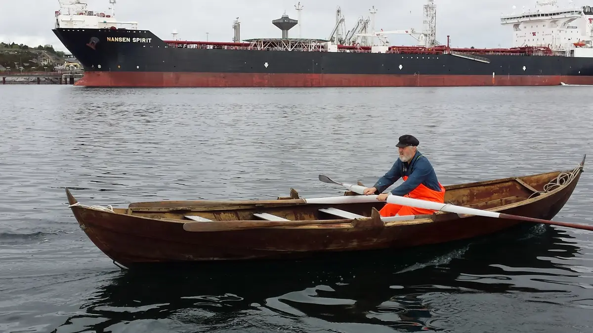 Ein mann i oransje oljehyre ror ein færing. I bakgrunnen ligg ein tankbåt ved kai utanfor Sture oljeterminal.