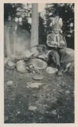 En gutt sitter ute i skogen ved et bål,  han har på seg et k