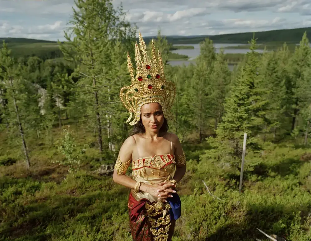 En kvinne kledd i gullfargede klær og stor krone står i en barskog. 