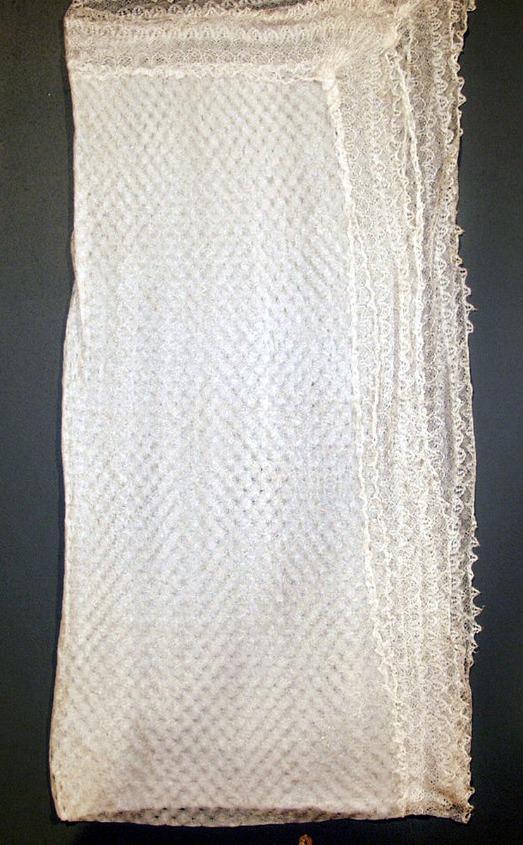 Sjal laget av tynt, strikket stoff i silke, lagt dobbelt. Påsydd bord med kniplede blonder, også av silke.