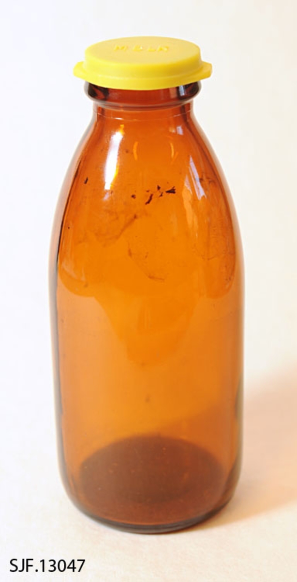 Melkeflaske bestående av flaske og lokk. Flaska er lagd av brunlig glass. Den er er 18,3 centimeter og har en sylindrisk kropp med 7,6 centimeters diameter. 11-12 centimeter ovenfor standflata skrår flaska slakt innover mot en krage rundt munningen. Her er diameteren (utvendig) 4,2 centimeter. I bunnen av flaska er det noen symboler i opphøyet relieff, noe som kan se ut som en sammensmeltning av bokstavene «M» og «G» (som indikerer at dette er et produkt fra Moss Glassverk), samt tallene «8» og «65». Flaska kan romme et væskevolum på 5 desiliter. Melk ble levert i slike flasker, med en foliekapsel over flaskemunningen. For å kunne lagre rester etter at foliekapselen var brutt hadde husholdningene gjerne en del plastdeksler som kunne gjenbrukes hver gang ei flaske med melkerester skulle settes til side i påvente av at resten av innholdet skulle konsumeres.  Denne flaska ble avlevert til museet med et deksel
av gulfarget plast. Denne komponenten har innskriften «MELK» i opphøyet relieff på toppen, og den har to «flipper» for å kunne vippe av lokket.