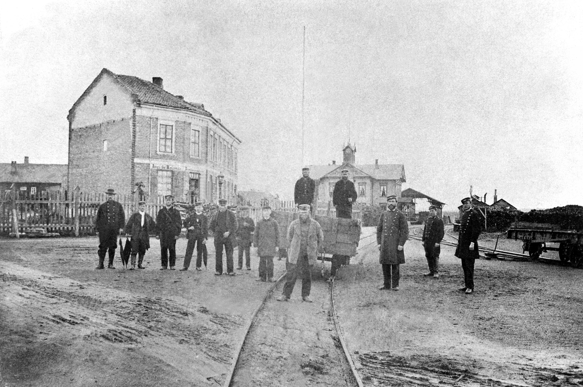 Hamar jernbanestasjon, stasjonsbygning nr. 1 fra 1862, Grundsetbanen, Kluges Hotell til venstre kalt "Jernbanehotellet", gruppe 13 personer. Banemester Ralph King står fremst midt i bildet, stasjonsmester Chr. Lund og kontorist Hans Eriksen står til høyre.
