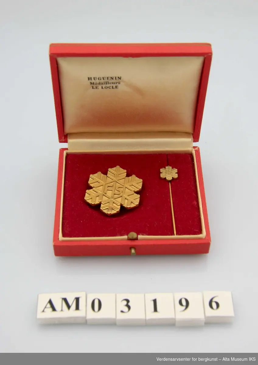 Etui med FIS-merker: 1 på stud (pin) og 1 på nål
Snøkrystallform med initialene FIS (Disse er miniatyrer av medalje AM.03195)
Etuiet er rødt og gult, 9 x 7,5 x 2 cm