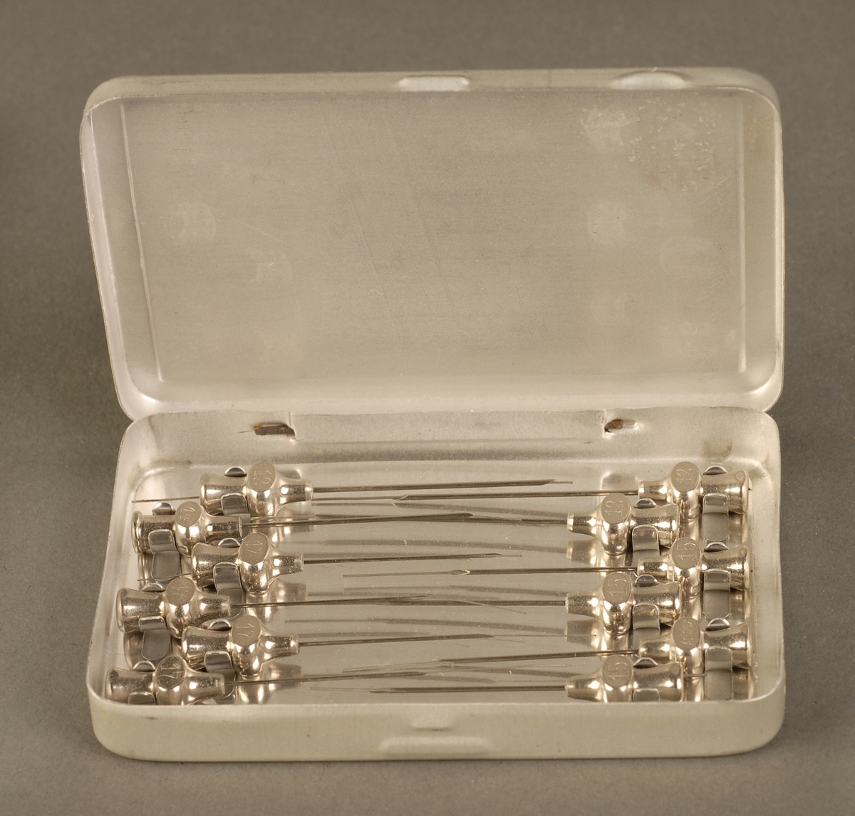 Rektangulær metalleske med hengslet lokk, inneholder 12 kanyler til injeksjonssprøyte