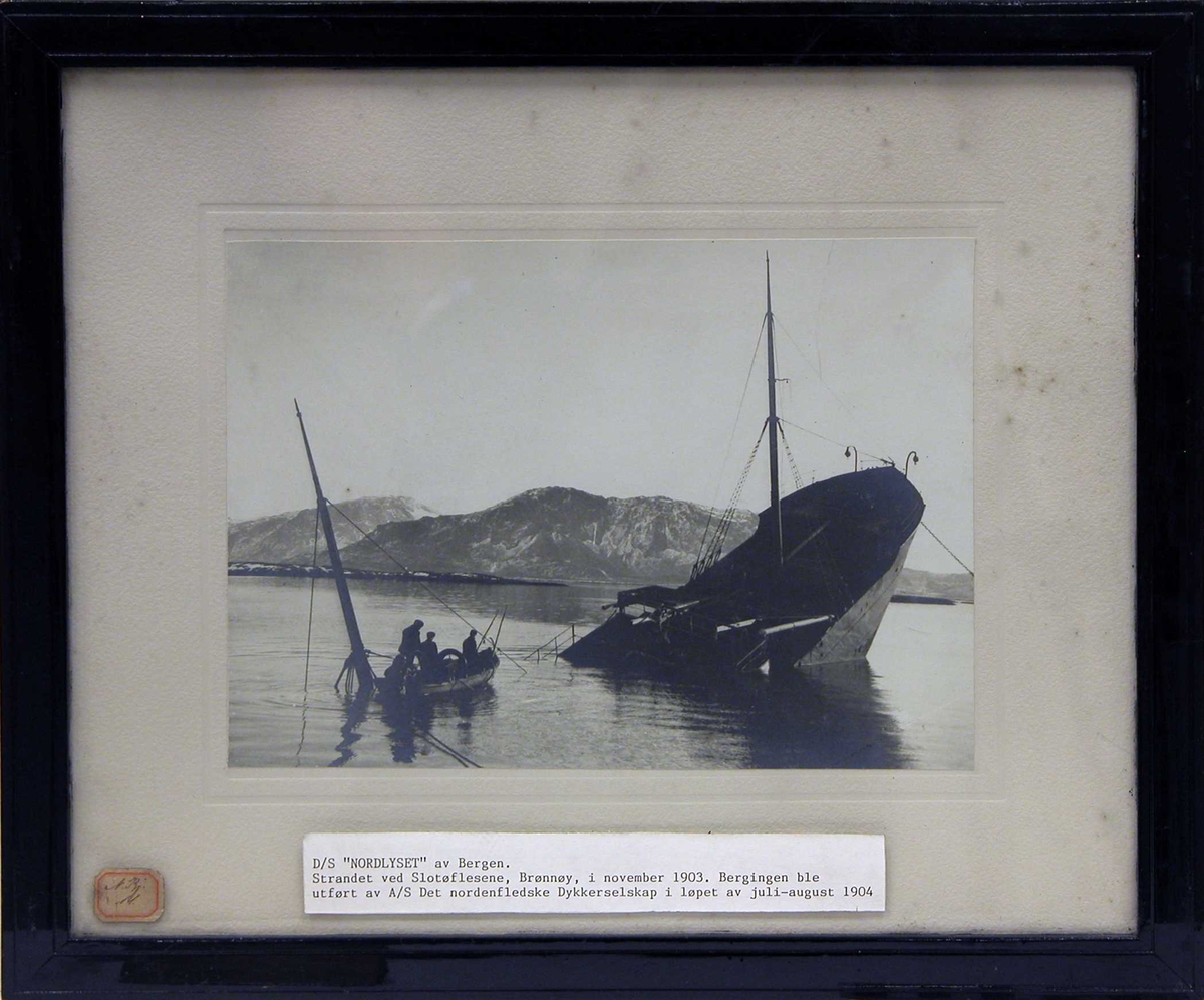 Fotografi av det havarerte skipet S/S "Nordlyset" av Bergen. Vi ser akterenden stikke opp. Slotøflesene ved Brønnøy november 1903.