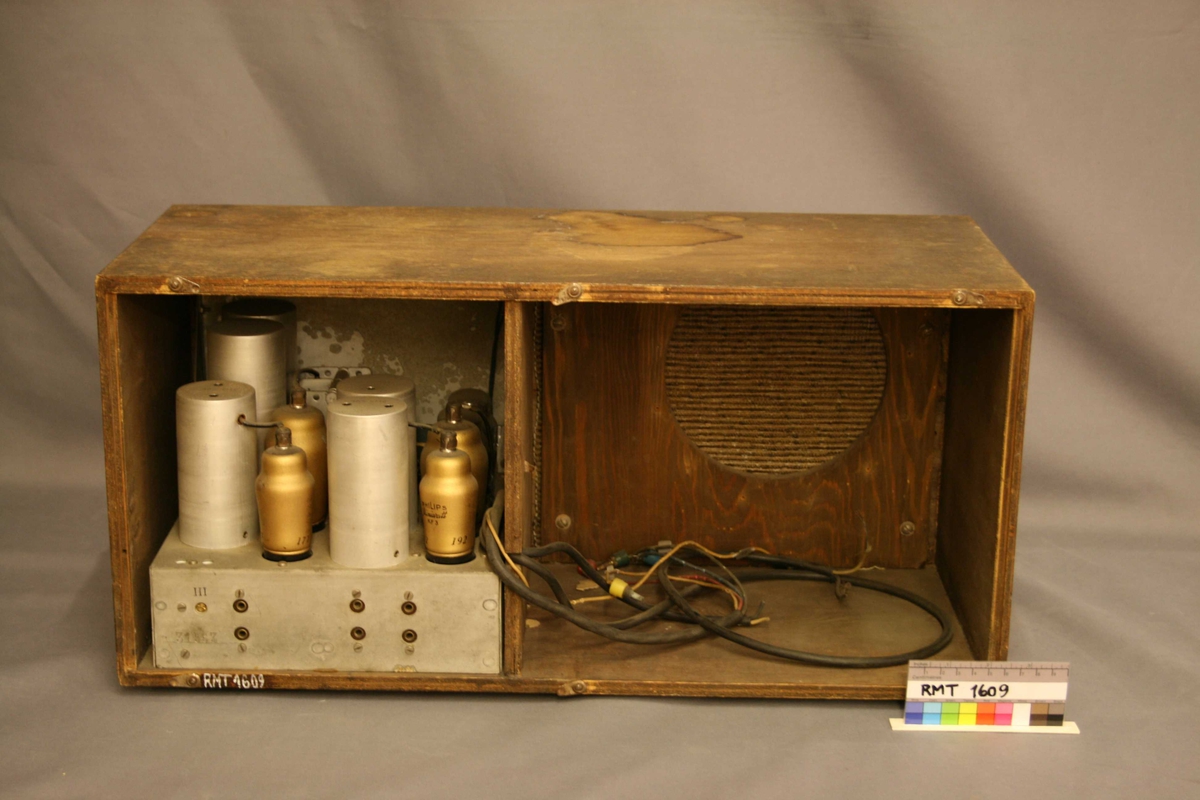 Rektangulær brun radio. Forside: skala med fem funksjons- og søkeknapper. Høyttaler med trekk av tekstil. Bakside: hele bakplaten mangler. 