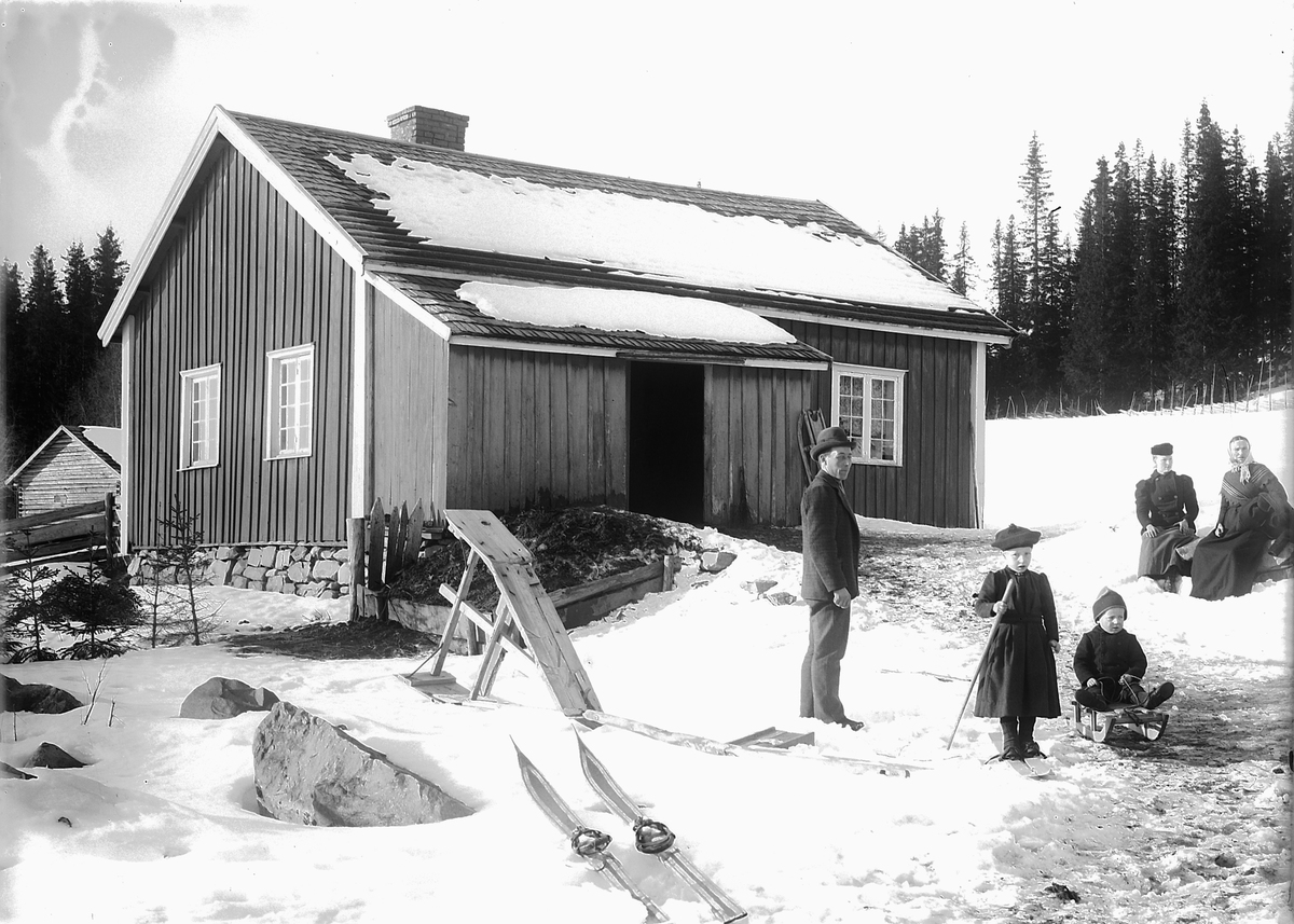 Ukjent familie foran et hus, ei jente på ski og en gutt på kjelke. 
