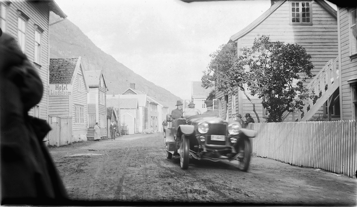 To menn i en bil kjører gjennom ei gate. Et skilt med Hotel Lindström til venstre. Lærdal i Sogn og Fjordane? Ola Hakkestad (1877-1922) og hans Minerva bil E1448.