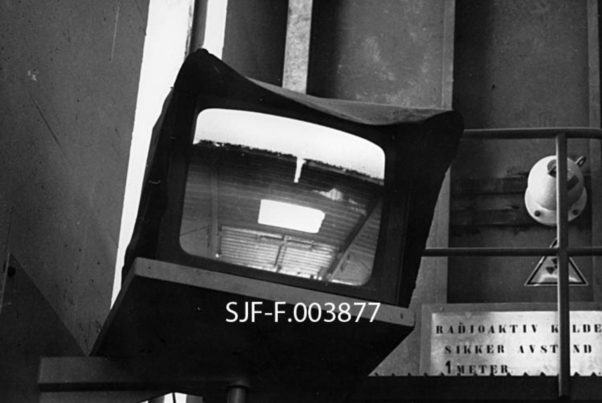 Monitor for overvåking av produksjon og transport av flis ved Geithus Bruk på Modum i Buskerud.  Fotografiet er tatt i oktober 1967, forholdsvis kort tid etter at produksjonen i dette anlegget var satt i regulær drift.  Vi ser en monitor (TV-skjerm som er plassert på ei hylle i produksjonslokalet.  Monitoren er overdekt med et mørkt materiale som stikker noe fram i overkant, antakelig for å forebygge reflekser fra omkringliggende lyskilder.  Bildet som ble vist på skjermen da dette fotografiet ble tatt later til å være fra en maskin.  Bak hylla med monitoren aner vi en plattform med rekkverk av jernrør.  Vi ser også en plakat som varsler om er radioaktivt objekt, og oppfordrer til å holde en minsteavstand på en meter.  Om det var monitoren som ble ansett for å medføre strålingsfare vet vi ikke. 