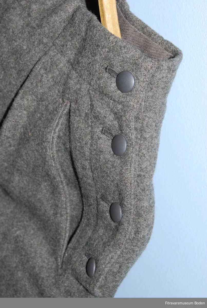 Av gråbrungrönt ylletyg och knäppta på sidorna med fyra grå släta knappar modell mindre. Två snedställda fickor. Byxbenen har slejf med spänne nertill. Storlek 42.