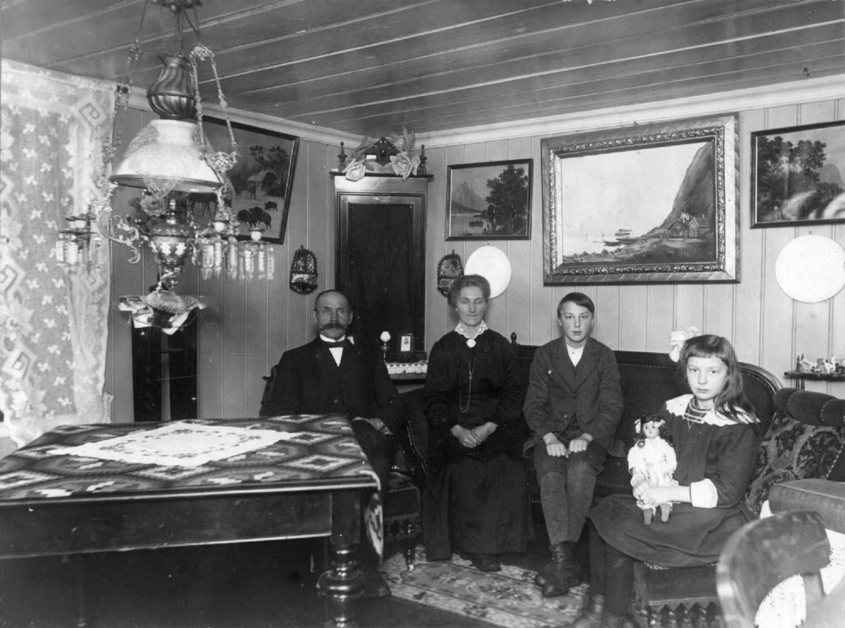 Stueinteriør, Ladderud gård på Eidsvoll, ca. 1915-1918. Huset ble bygget i 1842. Parafinlampe i taket, malerier på veggene. Fra venstre ekteparet Ole (gårdbruker) og Kaia Olsen Ladderud med barna Paul og Margit.