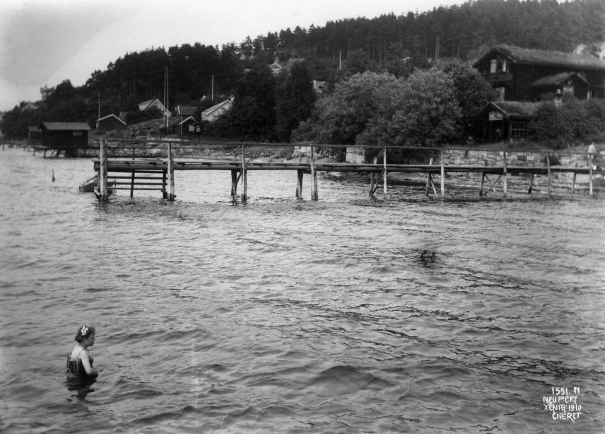 Thorkelbyen, Drøbak, Frogn, Akershus 1910. Hus og brygger langs stranda. Jente bader.
