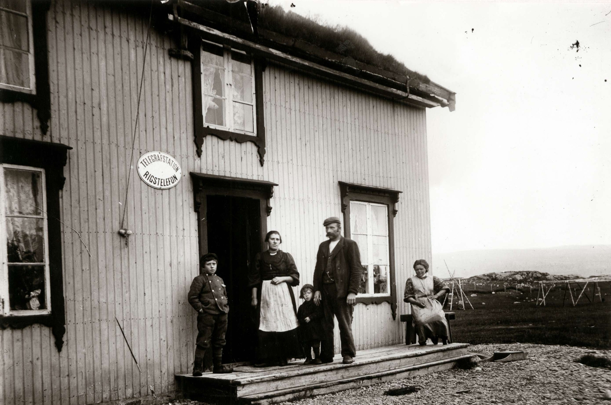 Persongruppe (5) foran telegrafstasjon, ant. Øst-Finnmark, trebygning i 2 etasjer. Skilt på veggen anviser også sentral for rikstelefon.
Del av serie fra en forskningsreise i Øst-Finnmark 1909.
