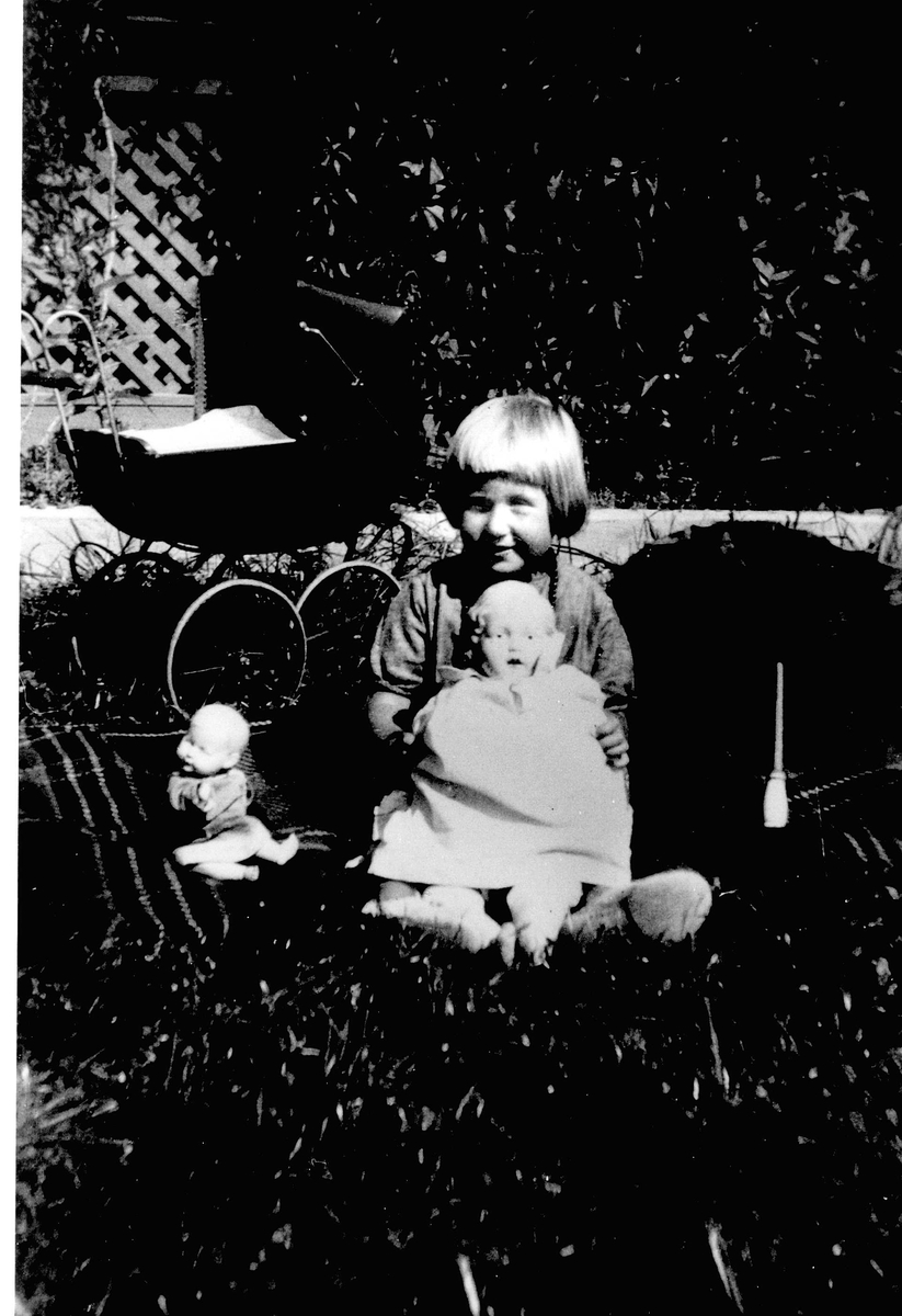 Pike med dukke og dukkevogn i hage. Jorunn Fossberg  i hagen til huset familien leide på Nærsnes, Røyken, Buskerud sommerne 1929-36. Bildet er tatt ca.1931. Dukkevognen ble da oppfattet som gammeldags, parasollen var rød med blomster.
Fra Jorunn Fossberg familiealbum.
