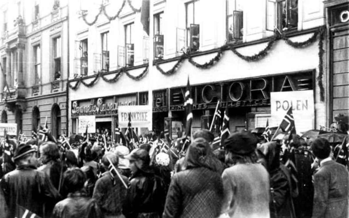 Fra Oslo under fredsdagene i 1945.
Publikum med plakater fra Holland, Belgia og Polen langs fortauet på Karl Johans gate, foran Bennetts Reisebyrå og Victoria billettservice.