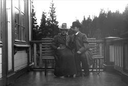 Par sittende på benk på veranda. Avfotografert(?)