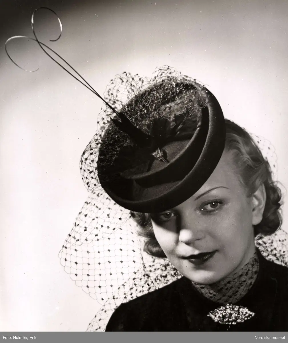 Hattmode visas på Nordiska Kompaniet 1941. kvinnlig modell i liten rund hatt med flor och upprättstående dekoration. I halsen bär hon en brosch med stenar. 