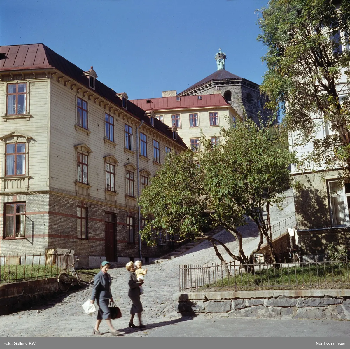Göteborg. Trähus, kullerstensgata. Kvinnor och ett barn i förgrunden. Skansen Kronan.