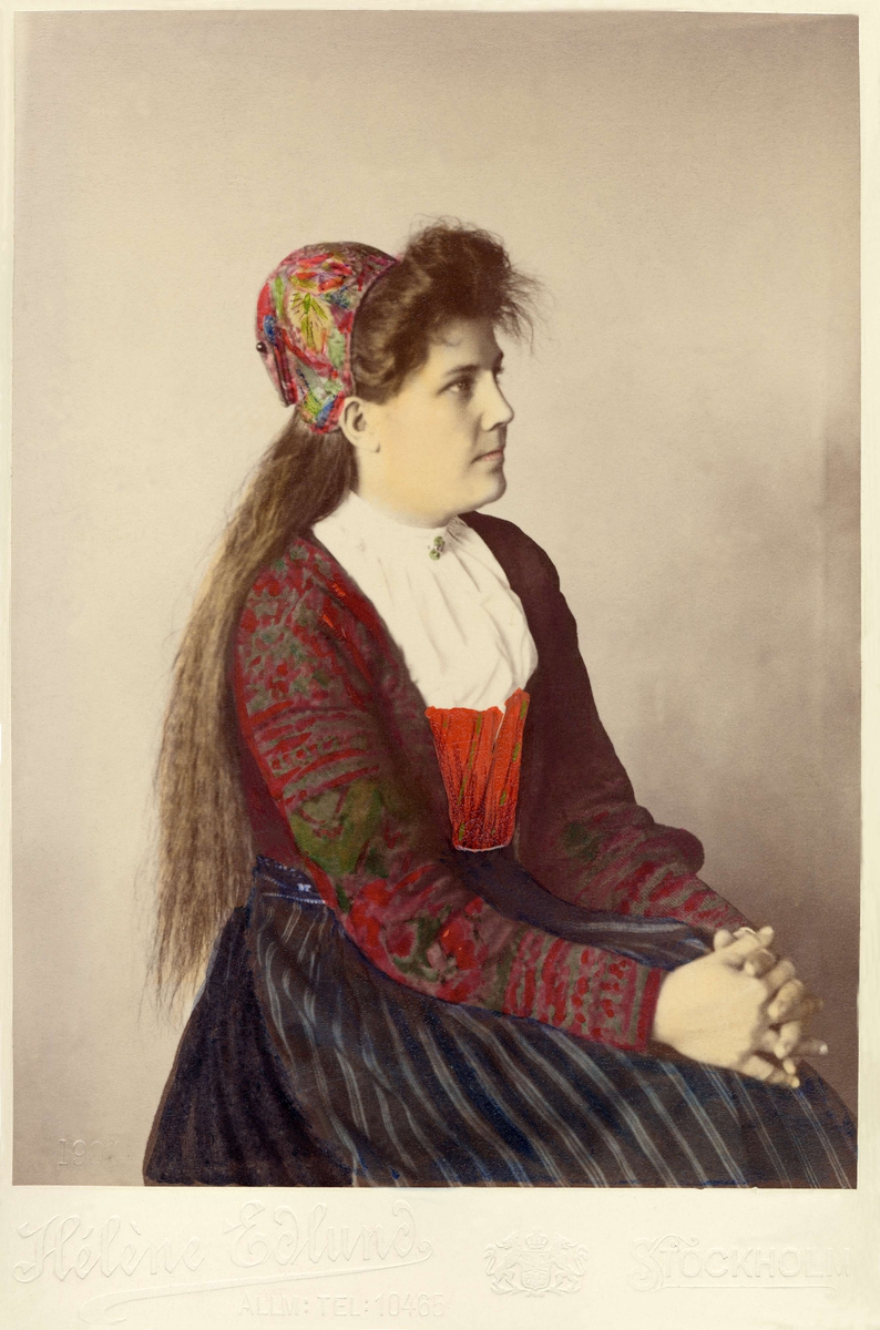 Foto, taget snett framifrån, på en ung kvinna i dräkt från Delsbo, Hälsingland i stickad tröja och kattunmössa s.k. påsluva