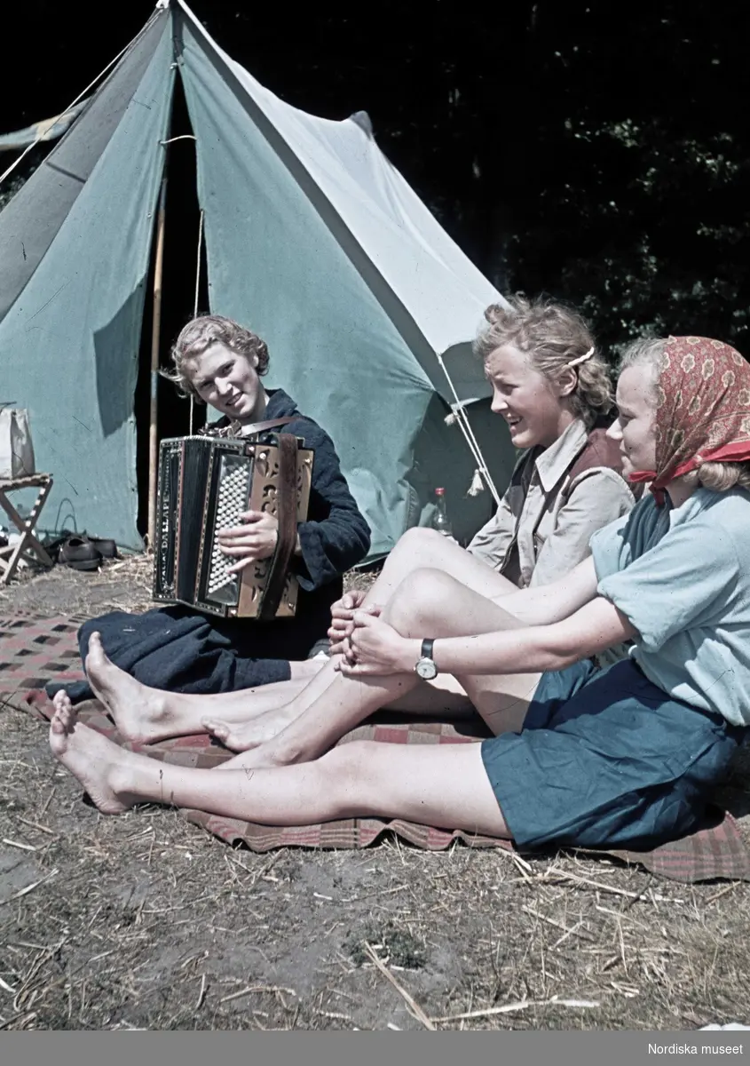 Tre flickor sitter på en filt utanför ett tält. Den ena flickan spelar dragspel.