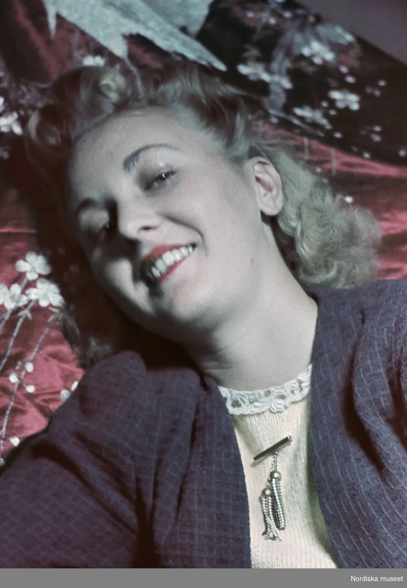 Porträtt av blond kvinna med rött läppstift, stickad blus med en brosch.