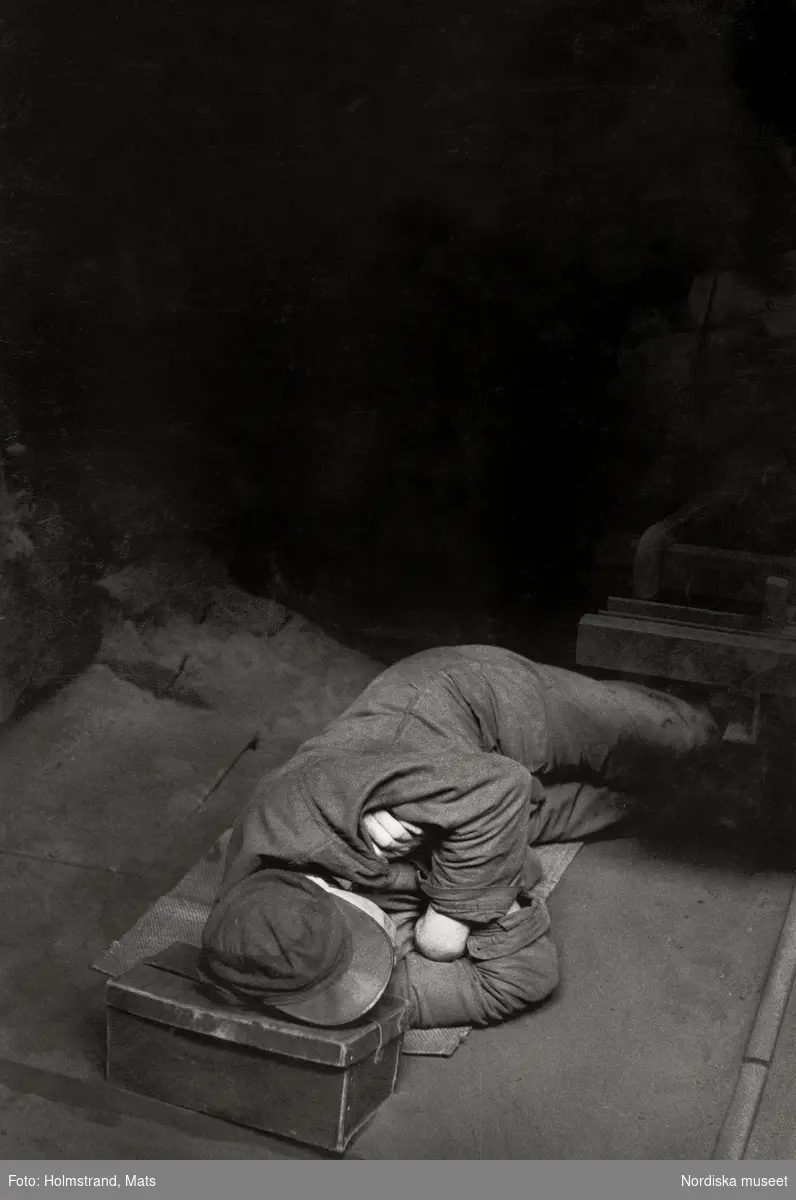 Man iförd arbetsläder, overall och keps, ligger på golvet och vilar. Som huvudkudde har han en unicabox. Interiör från gjuteriet på Albin Motor AB i Kristinehamn, 1956.
