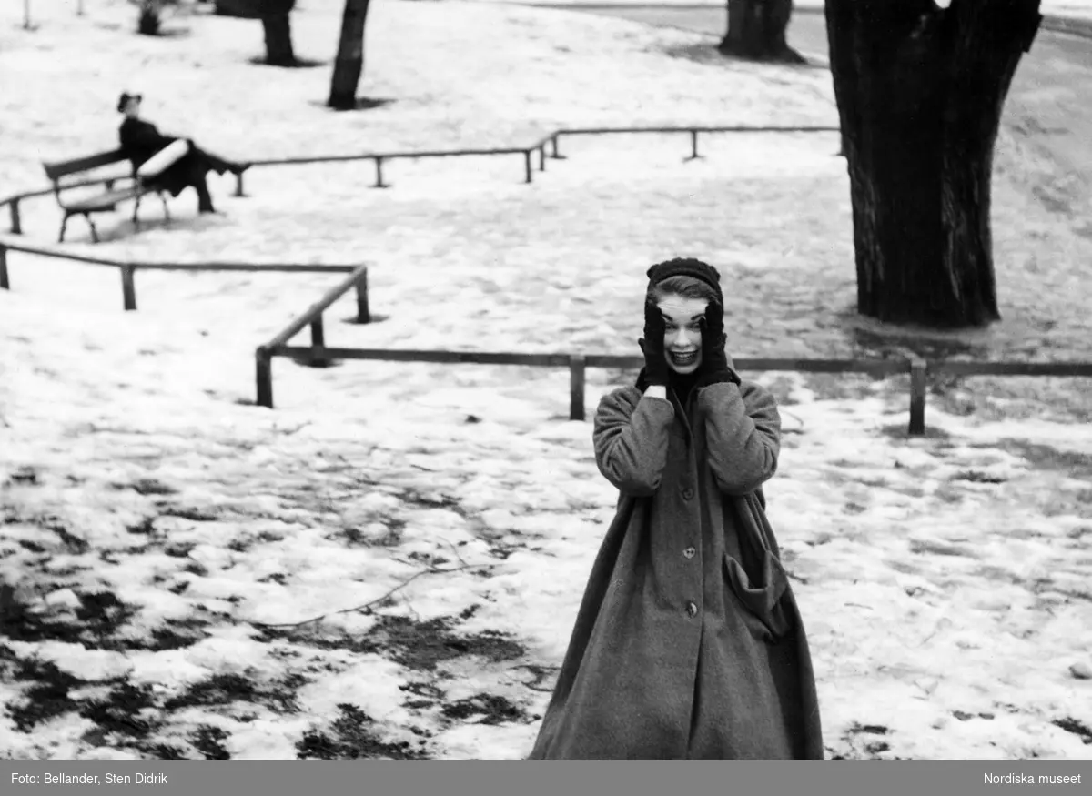 Vinter. Modebild. Kvinnlig modell trycker händerna mot kinderna. Hon står i en park med snö. I bakgrunden sitter en man med hatt och rock på en bänk. Stockholm.