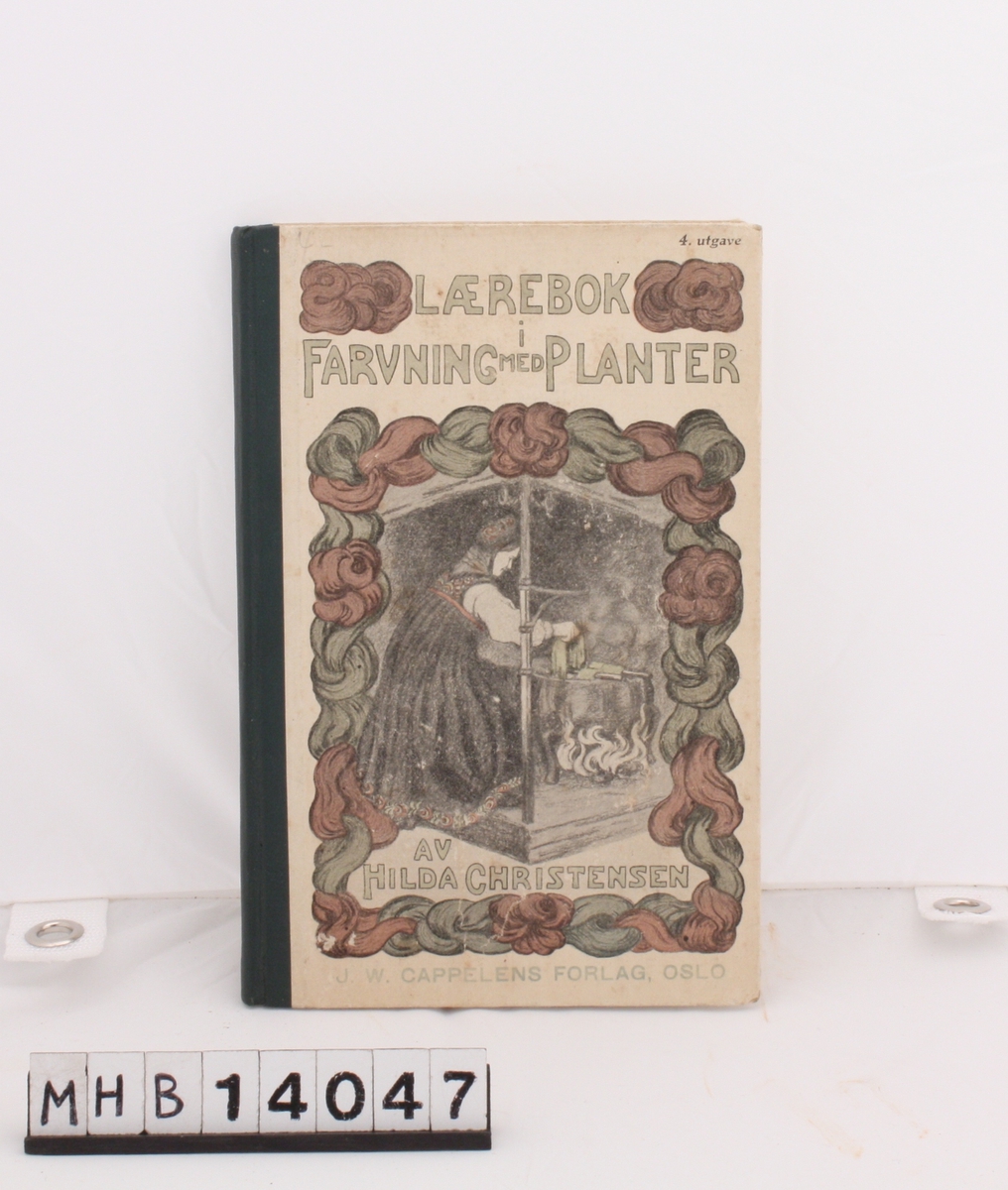Rektangulær bok med pappinnbinding og grønn rygg. På forsiden er en kvinne i bunad som plantefarger i jerngryte over åpen ild. Motivet er innrammet av røde og grønne ullhespler.