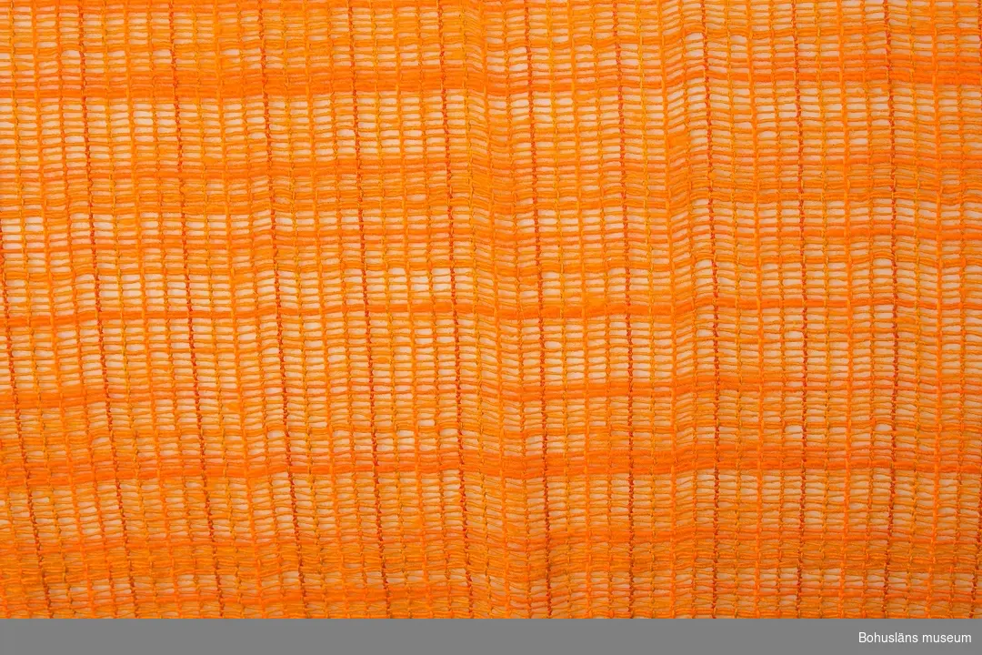 Glest vävd gardin (par) av syntetfiber i intensivt orange färg i något svagt varierande nyanser. En typisk färg för inredningsdetaljer från omkring mitten av 1960-talet till början av 1970-talet. Upptill tunnel för gardinstång. Nedtill 42 cm bred fåll, troligen tillkommen för att korta gardinerna.                                               
Personuppgifter och fakta kring samlingen se UM025572.