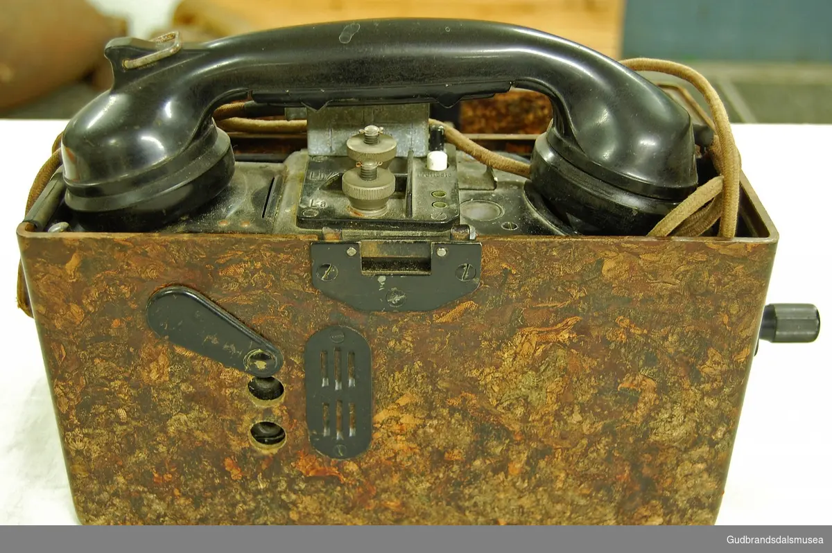 Svart telefon bygd inn i en brun, bærbar kasse. Rektangulær kasse med hengslet lokk. Med sveiv. Koblingsskjema med tysk tekst, alfabetkoder på tysk.