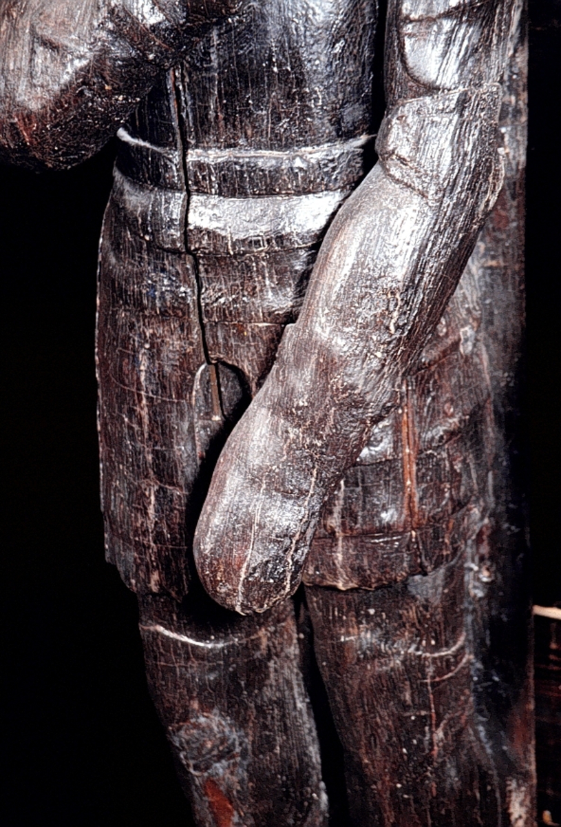 Skulptur av riddare i 1600-talsrustning med öppet visir.
Krigaren har kraftiga mustascher av typ "knävelborrar" och krokig näsa. Hakan är dold av hjälmen och fältbindeln draperad som en rund krage kring bröstets övre del. Rustningen är stiltrogen med sitt korta bröstharnesk med ansats till så kallad tapul, det vill säga den spetsformiga utbuktningen strax under bröstets mittpunkt, sina långa, mot knäna neddragna lårskört samt knä- och armbågsskydd.

Huvudet är något vridet åt höger. Höger arm hålls lyftad framför bröstet medan vänster är sträckt snett framför kroppen. Vänster ben är något böjt men kroppstyngden förefaller vara någorlunda jämnt fördelad på bägge benen. Främre delen av fötterna saknas. Fötterna har ursprungligen stuckit utanför den smala fotplattan eller sockelns framkant. Skulpturen har troligtvis varit utrustad med svärd eller lans, kanske också sköld.

Skulpturen är snidad i ett stycke så när som på armar och höftparti som är utförda separat och fastsatta med dymlingar och spikar. Även fötternas ytterändar har varit separat snidade. Baksidan är slät. Upptill är bakstycket snett uppdraget ovanför skulpturens huvud och bildar en kraftig huvudplatta. Fotändan är snedskuren.
Skulpturen är i mycket gott skick.

Text in English: Sculpture of a knight in seventeenth-century armour, with a "handlebar" type moustache and a crooked nose.
His chin is hidden by the helmet and the field badge which is draped round the upper part of his torso and neck like a collar. The armour is faithful to the style, with its short cuirass and breastplate tapered to a point and long tuille, and cubitieres and knee pieces.

The head, its face stern, is twisted slightly to the right. His right arm is held up and across the front of his chest, while the left falls in front of and against his body. The left leg is slightly bent, but the weight of his body appears to be evenly distributed over both legs. The front sections of the feet are missing but originally protruded over the front of the narrow base upon which the figure stands.
The sculpture is well preserved.