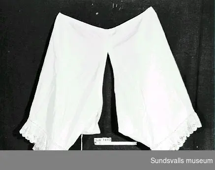 Benkläder av vit bomull med spets på benens kant. På framsidan är namnet 'Ella' broderat i kedjestygn.