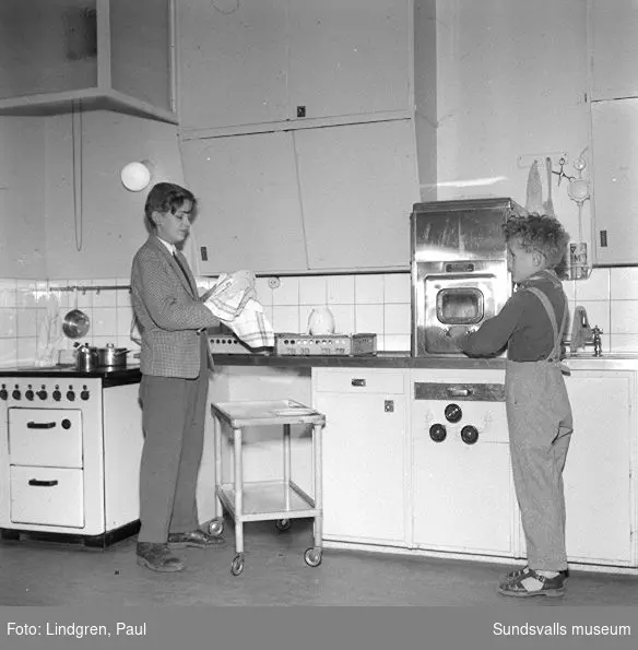 Fru Lindlöf i sitt kök, samt två pojkar som hjälper till med disken.