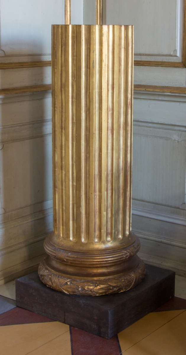 Piedestal av trä. Formad likt en kolonn på en rund bas. Kolonnen med profilerade, skurna och förgyllda kannelyrer. Basen med svarvad, skuren, skulpterad bandstav och lagerstav. Underst en kvadratisk sockel imitationsmålad likt sten.