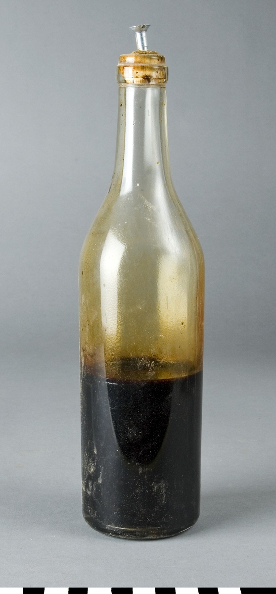 Flaska av ofärgat glas fylld till ungefär en trdjedel med okänd brun vätska. I korken som försluter flaskan sitter en skruv.