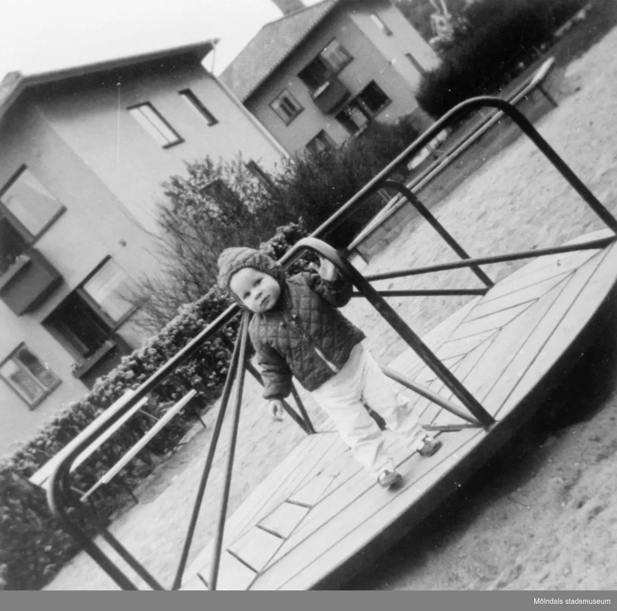 Ett litet barn ståendes i karusell i en lekpark, 1965. Brunnsgatan 4 och 6 syns i bakgrunden.