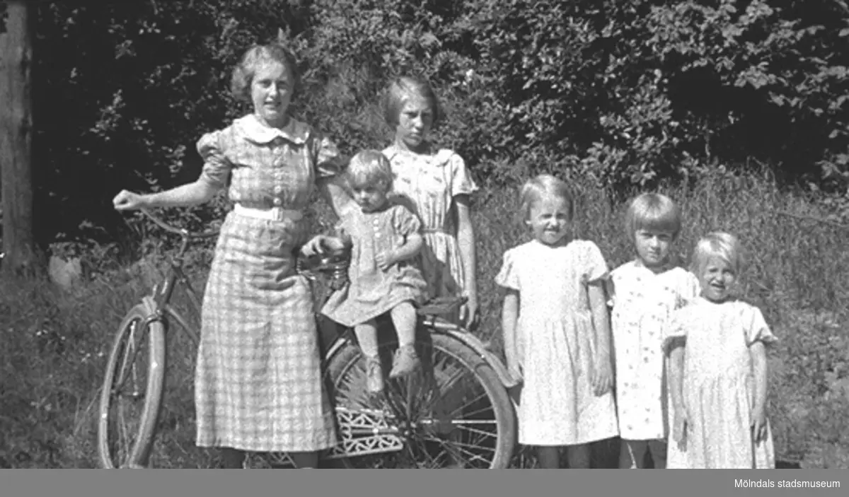 Barnen i familjen Karlsson står vid en cykel på Kvarnfallet 31, år 1936.  Från vänster: Astrid (gift Garthman), Rolf (på cykeln), Margit (gift Larsson), Birgit och Ingrid. Något utav barnen är okänt. Skänkt av sonen Alf Garthman som arbetade under många år på Mölndals stadsmuseum.