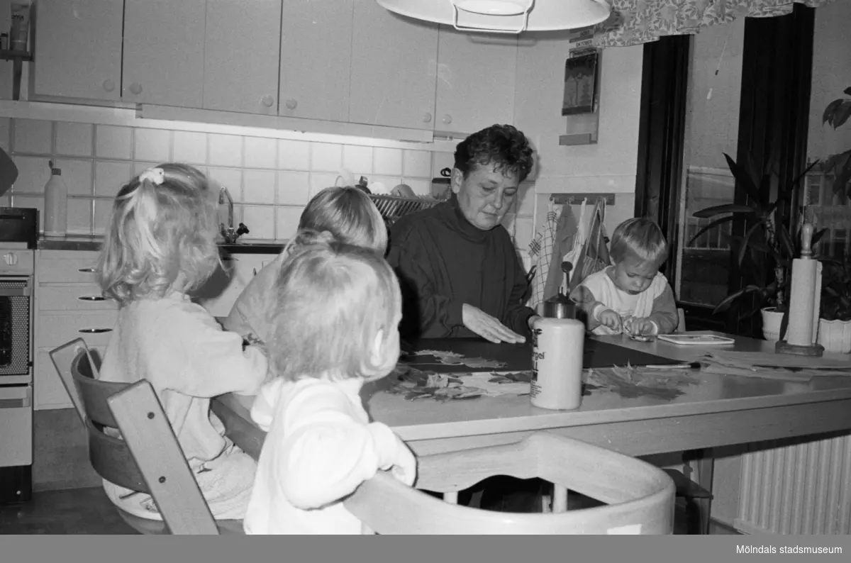 En kvinna och tre barn sitter runt ett bord. Ett utav barnen sitter på en trip trap stol och ett barn står bredvid en barnstol. De lägger pussel och pysslar, i köket på Katrinebergs daghem.
