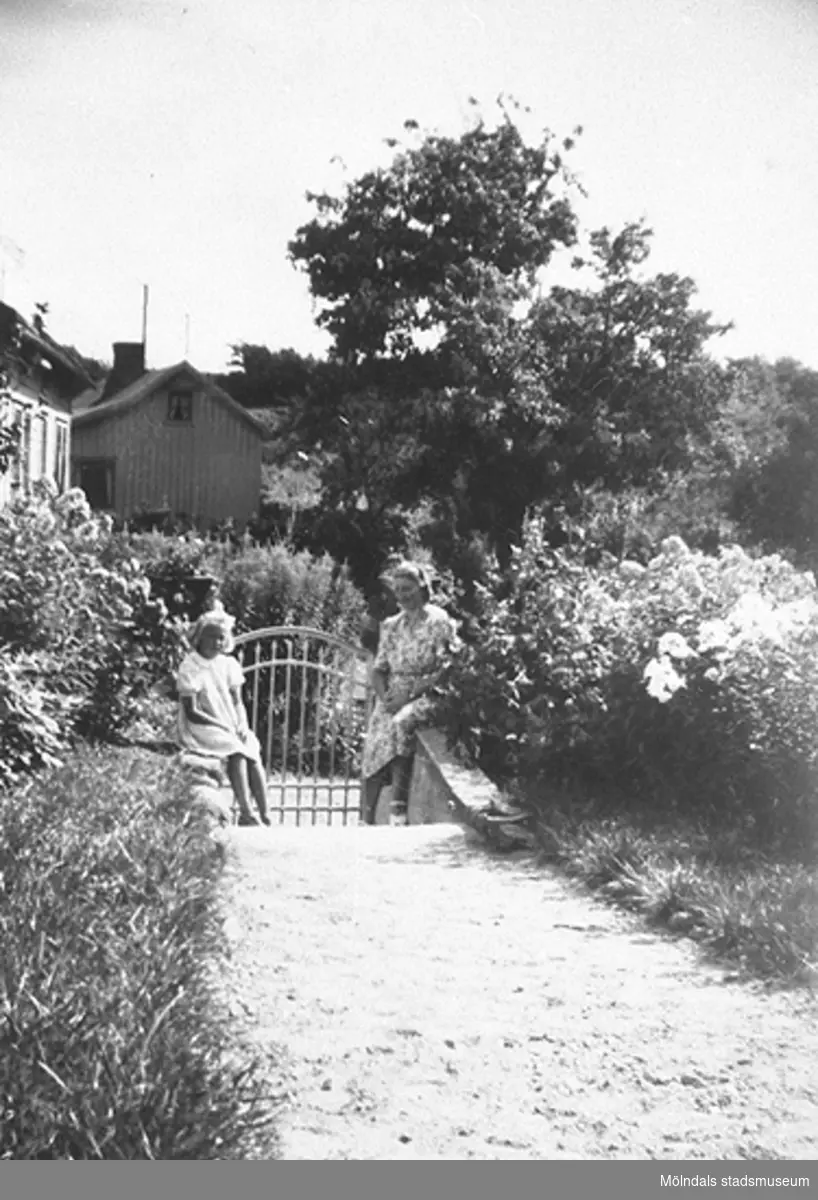 Sommardag på Kryssgatan i Mölndal i början av 1940-talet. Anna Larsson (född Hasselberg) med dottern Berit står vid grinden i husets trädgård.
