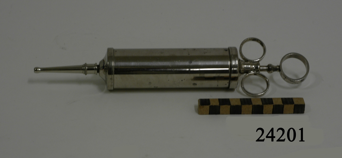 Cylinderformad spruta av metall med skruvlock,  metallkolv och tre fingerringar. Påskruvbar rak kanyl, 6 cm lång med olivformad spets. Märkt: "KIFA" SAMT "M".