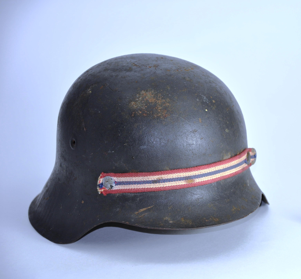 Hjelm i metall, polstret med skinn innvendig. Rem til å ha under haken. Hjelmen er opprinnelig tysk, M1943, men er "nasjonalisert" ved bortskrapning av tysk emblem og med påklinket bånd i norske farger.